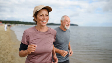 Active senior couple runners jogging outdoors on sandy beach by sea in early morning.
