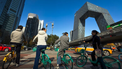 Cyclists Wait At An Intersection Near The CCTV Headquarters In Beijing