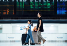 Smiling young Asian mother and daughter travelling together, they are carrying suitcases and walking against arrival departure board in airport terminal. Ready for a trip. Family travel and vacation concept