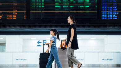 Smiling young Asian mother and daughter travelling together, they are carrying suitcases and walking against arrival departure board in airport terminal. Ready for a trip. Family travel and vacation concept