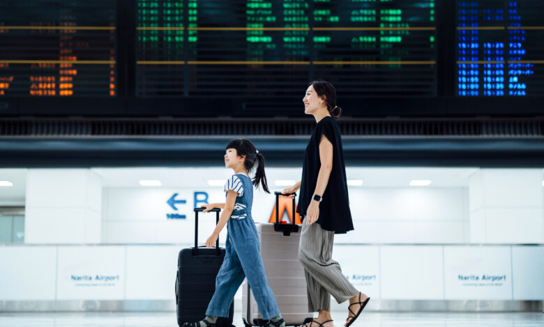 Smiling young Asian mother and daughter travelling together, they are carrying suitcases and walking against arrival departure board in airport terminal. Ready for a trip. Family travel and vacation concept