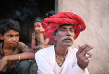 Senior Rabari man smoking a beedi sitting outside their house wearing a red turban looking at the camera