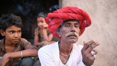 Senior Rabari man smoking a beedi sitting outside their house wearing a red turban looking at the camera