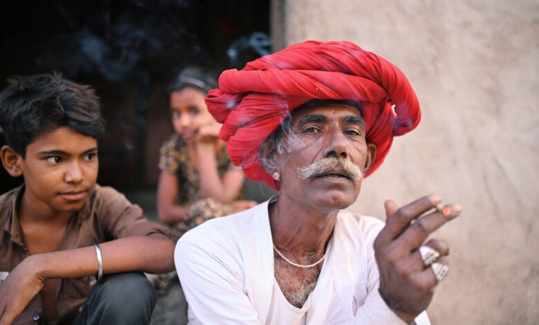 Senior Rabari man smoking a beedi sitting outside their house wearing a red turban looking at the camera